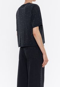 Černá košile s krátkým rukávem a rozptýlenými kovovými ozdobami na hladkém materiálu. Stylizováno s volnými černými kalhotami se standardními kapsami.