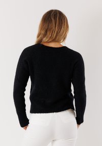 Maglione nero a maglia con maniche lunghe, tessuto testurizzato e scollatura rotonda, indossato su pantaloni bianchi, che mette in evidenza un design cropped.