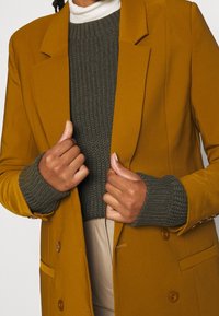 Veste moutarde jaune à coupe ajustée, portée sur un pull en maille vert foncé avec une texture côtelée et des manches longueur poignet.