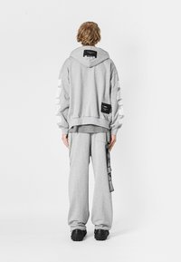 Sudadera gris con acentos de franjas blancas y etiqueta negra. Combinada con pantalones de chándal grises holgados que tienen un bolsillo lateral y detalles en negro.
