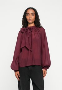 Femme aux cheveux bouclés portant un chemisier bordeaux à manches longues avec un grand nœud au niveau du col et un pantalon noir, sur fond blanc.