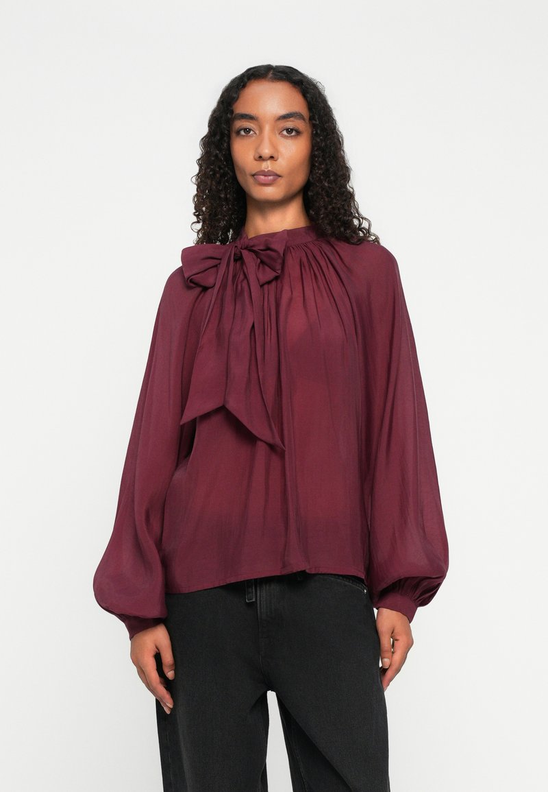 Femme aux cheveux bouclés portant un chemisier bordeaux à manches longues avec un grand nœud au niveau du col et un pantalon noir, sur fond blanc.