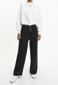 Sudadera blanca recortada con logo negro, combinada con pantalones negros anchos de chándal y zapatillas blancas con superficie texturizada.