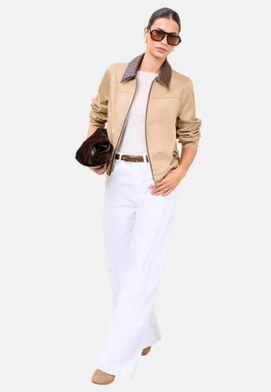 Vrouw met beige jas met donkerbruine kraag, witte broek, bruine riem, grote zonnebril, houdend een bruine hoed tegen een witte achtergrond.