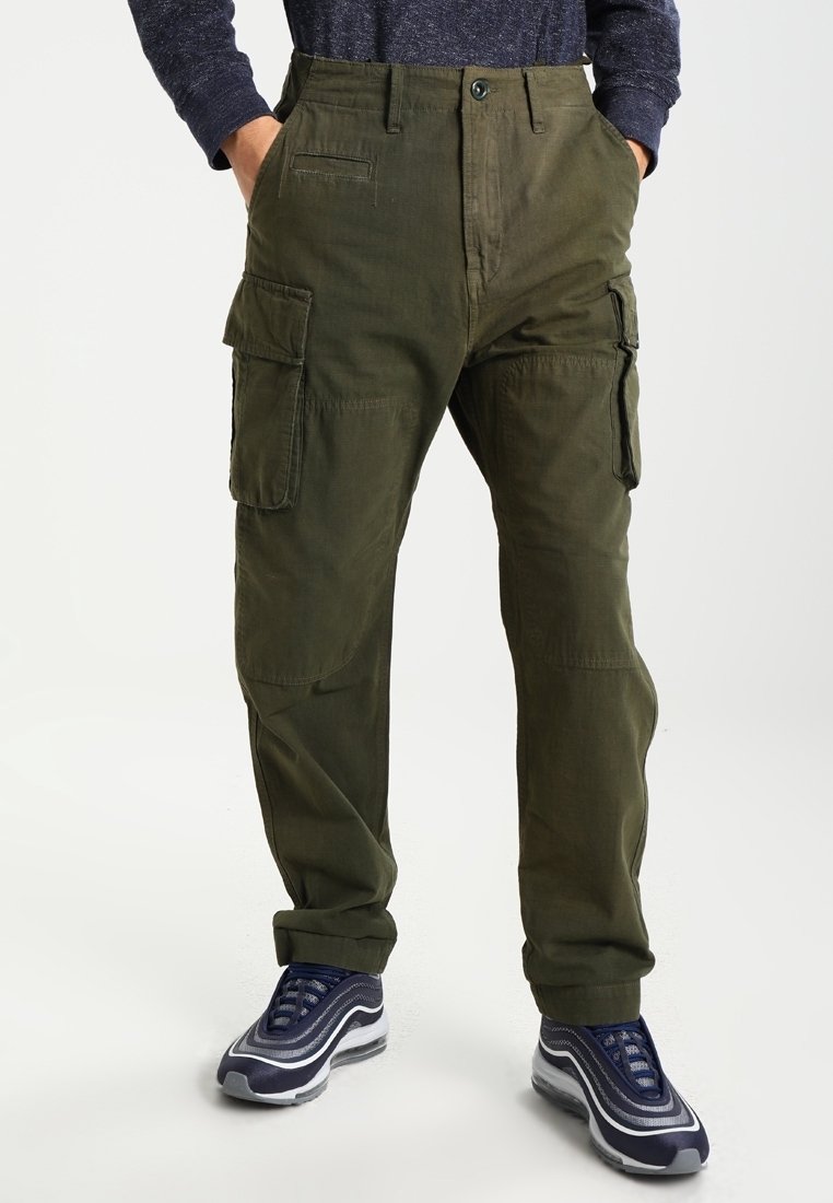 Cargo byxor i olivgrön, med flera sidofickor, en lös passform och ett lätt strukturerat bomullstyg. Bärs med marinblå sneakers.