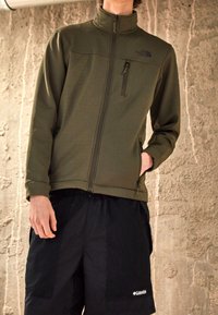 Olivově zelená zipová bunda s vysokým límcem a černým logem, kombinovaná s černými síťovanými šortkami s bočními kapsami a značkovými detaily.