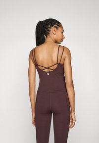 Brown sports tank top with thin straps, featuring a crisscross design at the back. Smooth texture and fitted silhouette, paired with matching leggings.