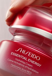 Roter Behälter der Shiseido Essential Energy Feuchtigkeitscreme mit silbernen Akzenten. Glatte Textur, das Produkt ist sichtbar, eine Hand greift den Deckel.