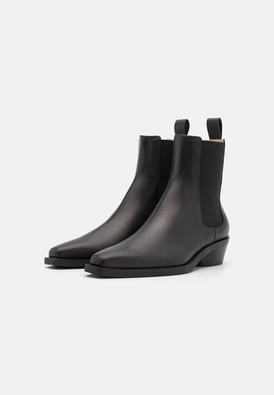 Botas de tobillo de cuero negro con un diseño elegante, paneles laterales elásticos, punta afilada y un tacón bajo tipo cuña. Textura limpia y suave.