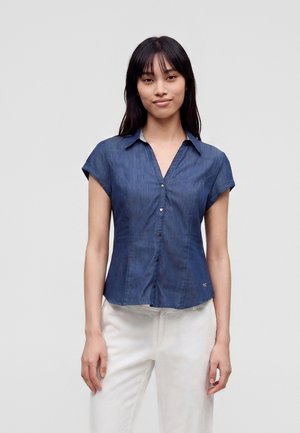 Jeune femme aux longs cheveux foncés, portant une chemise bleue à manches courtes boutonnée et un pantalon blanc, debout devant un fond clair uni.