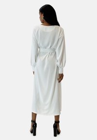 Robe blanche à manches longues en tissu lisse, avec une taille cintrée par une ceinture, tombant jusqu'au milieu du mollet. Portée avec des chaussures à talons noires.