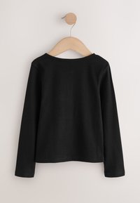 Chemise noire à manches longues en tissu côtelé, avec un col large et un ourlet droit. Suspendue sur un cintre en bois contre un fond neutre.