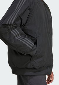 Casaco bomber preto em tecido suave e leve. Apresenta três riscas escuras em cada manga e punhos e barra canelados. Inclui bolsos com fecho.