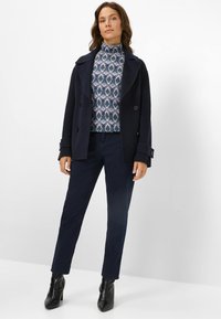 Cappotto di lana navy doppiopetto sopra un top con colletto alto a fantasia, abbinato a pantaloni scuri e stivali neri con tacco alto.