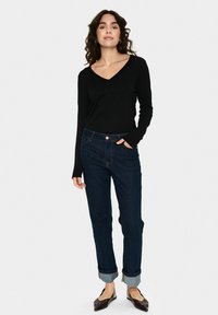 Fekete, hosszú ujjú V-nyakú felső, sötétkék, magas derekú farmernadrággal párosítva, bokán felhajtva, és barna lapos cipőkkel.