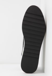 Semelle de chaussure en caoutchouc noir avec des rainures horizontales et un talon légèrement surélevé. Surface lisse avec des bords arrondis et un détail accentué blanc.