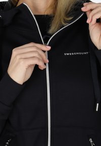 Marineblå zip-up sweatshirt med sølvlynlås og detaljer. Har hætte med snøre og logoet "SWEDEMOUNT" på brystet, glat tekstur.