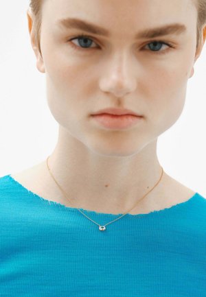 Collar de oro con un pequeño colgante de cristal, llevado por un modelo en un primer plano, luciendo una blusa texturizada de un brillante color azul.
