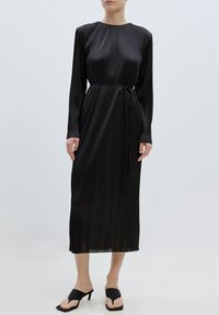 Schwarzes, plissiertes Midikleid mit langen Ärmeln, rundem Ausschnitt und Bindegürtel. Kombiniert mit schwarzen offenen High Heels. Glatte, glänzende Textur.
