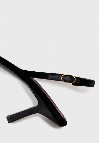 Svart sammet högklackad slingback sko med ett tunt band och en liten guldbuckla på vit bakgrund.