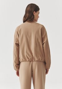 Hnědá bombarďácká bunda s uvolněným střihem, elastickým pasem a mírně zúženými rukávy. Hladká textura, minimalistický design.