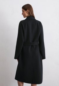 Long black wool coat with a belted waist, wide lapels, and dropped shoulders. The back features a simple seam detail and a smooth texture.
