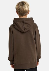Barn med kort, lyst hår som vender bort, iført en brun hettegenser med hette, mot en enkel hvit bakgrunn.