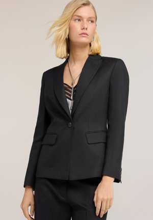 Blonde Frau trägt schwarzen, maßgeschneiderten Blazer und passende Hose, mit silberner Halskette und darunter einem schwarzgestreiften Top.