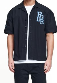 Camisa de botones negra de manga corta con cuello, que presenta un logo bordado en azul "PGDR" en el lado izquierdo del pecho y una camiseta blanca debajo.