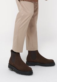 Brązowe zamszowe botki Chelsea z elastycznymi panelami bocznymi, grubą czarną gumową podeszwą i smukłą sylwetką idealną do codziennego noszenia.