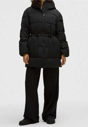 Personne portant un manteau noir matelassé à capuche avec poches avant, assorti à un pantalon noir ample et des chaussures noires sur un fond gris uni.