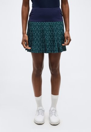 Personne portant un haut bleu marine, une jupe plissée verte à motif, des chaussettes blanches et des chaussures blanches à lacets sur un fond clair uni.