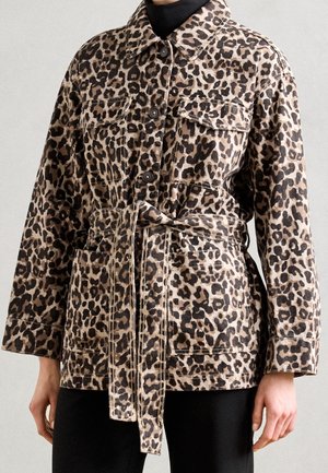 Kobieta w zapinanej na guziki kurtce w panterkę z paskiem i przednimi kieszeniami, nosząca czarne spodnie i czarny golf.