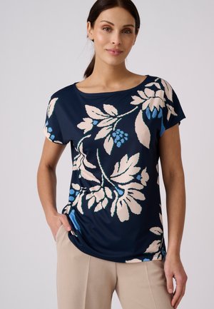 Mujer con cabello oscuro, con una camiseta azul marino de manga corta con estampado floral beige y azul, lleva pantalones beige claro, de pie con una mano en el bolsillo.