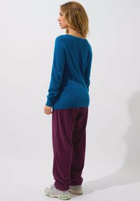 Blauer Pullover mit langen Ärmeln und weitem Ausschnitt, kombiniert mit lila Cordhosen. Das Model steht seitlich vor einem neutralen Hintergrund.