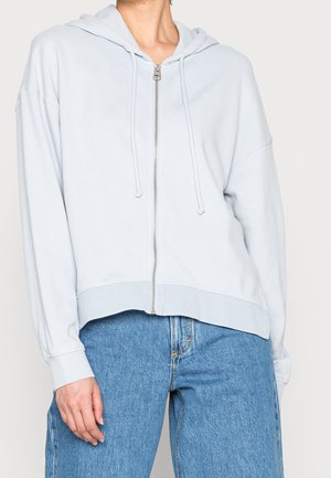 Personne portant un sweat à capuche gris clair zippé avec cordons, associé à un jean bleu taille haute, debout devant un fond uni.