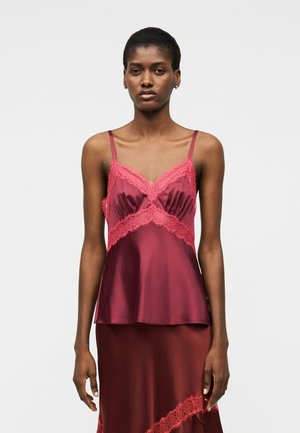 Kobieta ubrana w bordowy satynowy top bez rękawów z różową koronkową lamówką oraz dopasowaną satynową spódnicę na jednolitym szarym tle.