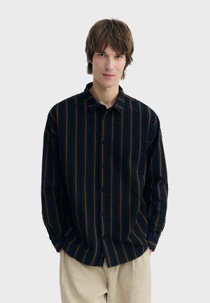 Jonge man met bruin haar die een zwart overhemd met verticale oranje strepen en beige broek draagt, staand tegen een effen achtergrond.