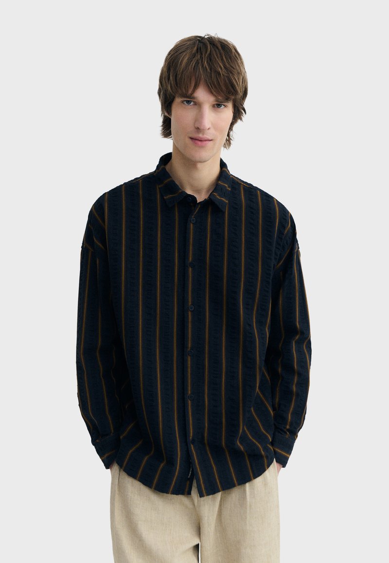 Junger Mann mit braunen Haaren, der ein schwarzes Hemd mit vertikalen orangefarbenen Streifen und beige Hosen trägt, vor einem schlichten Hintergrund stehend.