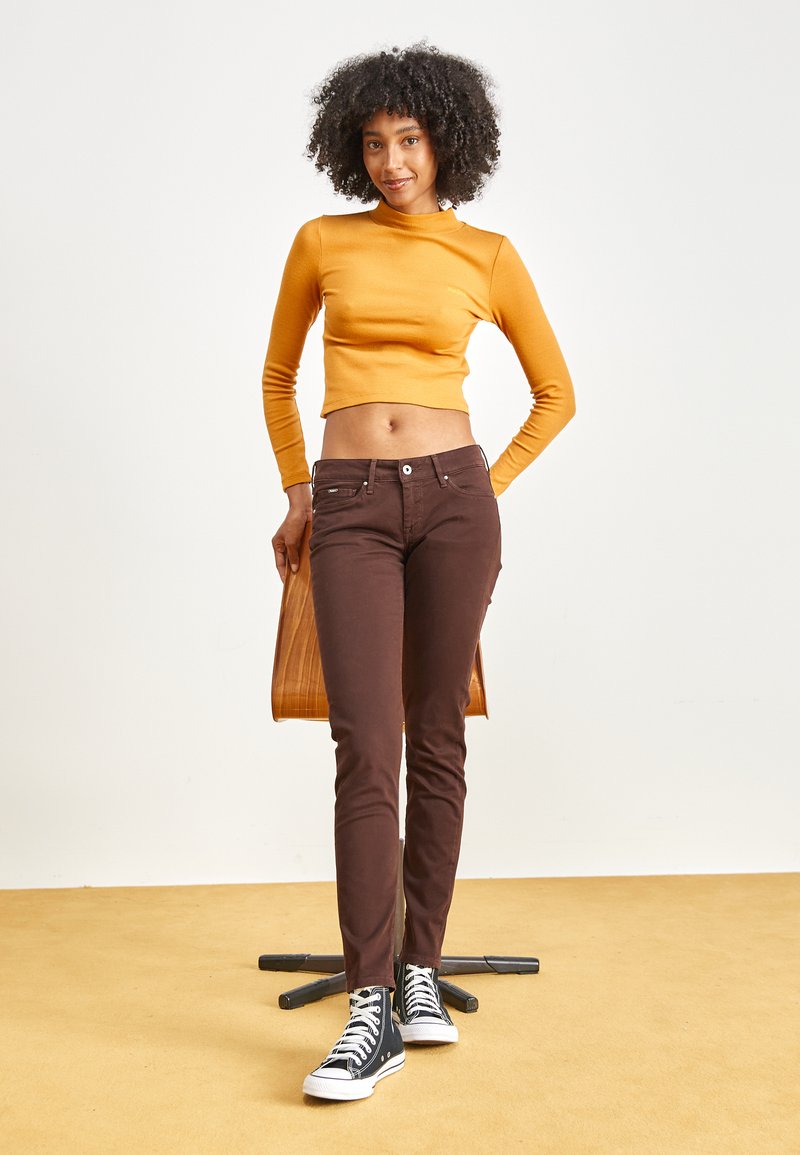 Braune Slim-Fit-Jeans, gerippetes senfgelbes Cropped-Top und schwarze High-Top-Sneaker; das Model steht mit den Händen in den Hüften vor einem einfarbigen Hintergrund.