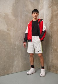 Giacca a vento rossa e nera, maglietta nera, pantaloni corti bianchi con logo Nike e scarpe da ginnastica bianche con dettagli rossi. Texture lisce e sportive.