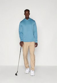 Ljusblå långärmad hoodie, khakifärgade byxor och vita sneakers. Står med en golfklubba. Enkel design utan mönster eller detaljer.