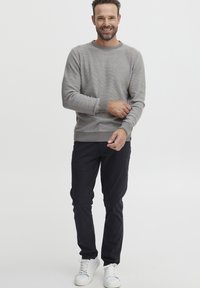 Maglione grigio a maglia con polsini e orlo a coste, abbinato a pantaloni neri e sneaker bianche. Il modello si trova di fronte a uno sfondo neutro.