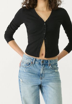 Zwarte geribde cardigan met V-hals en lange mouwen, voorzien van een knoopsluiting en een cropped ontwerp, gecombineerd met lichtblauwe distressed jeans.
