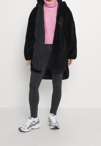Abrigo de felpa negro de gran tamaño con capucha, llevado sobre un suéter de punto rosa. Combinado con jeans skinny negros y zapatillas deportivas plateadas.