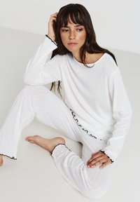 Un haut à manches longues côtelé blanc et un pantalon assorti présentent des accents froncés noirs aux poignets et à l'ourlet, le tout sur un fond neutre.