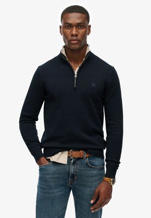 Navy zip-neck sweater made of soft fabric, featuring a contrasting collar and a discreet logo on the chest. Worn with blue jeans and brown belt.