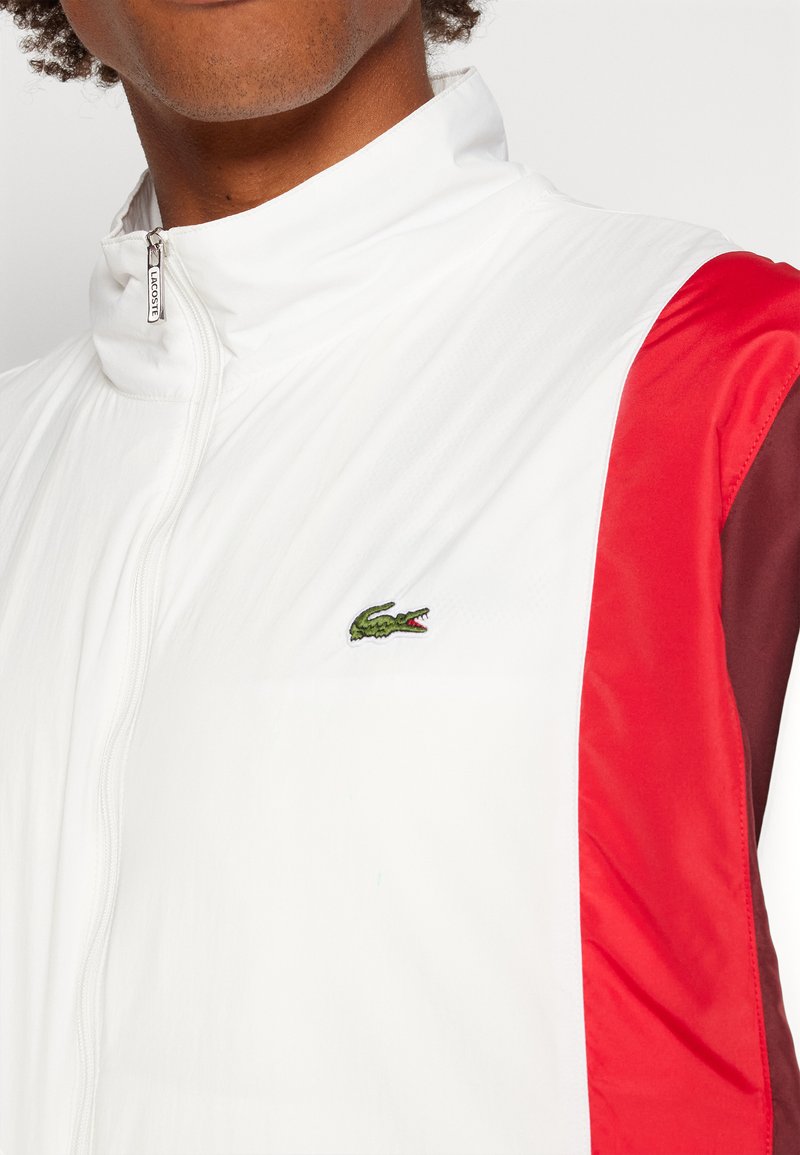 Veste blanche avec des accents rouges et marron, col haut, fermeture éclair, et logo de crocodile brodé en vert sur la poitrine gauche. Texture lisse.