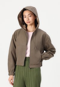 Chaqueta con capucha marrón de cremallera, con puños elásticos, de corte relajado y dos bolsillos frontales, combinada con una blusa rosa claro y pantalones verdes plisados.