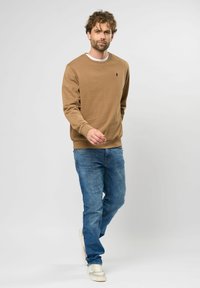 Ruskea huppari, jossa on pyöreä kaula-aukko ja ribbaatut hihansuut, yhdistettynä sinisiin farkkuihin ja valkoisiin lenkkareihin, kulkevalla miehellä vaaleaa taustaa vasten.